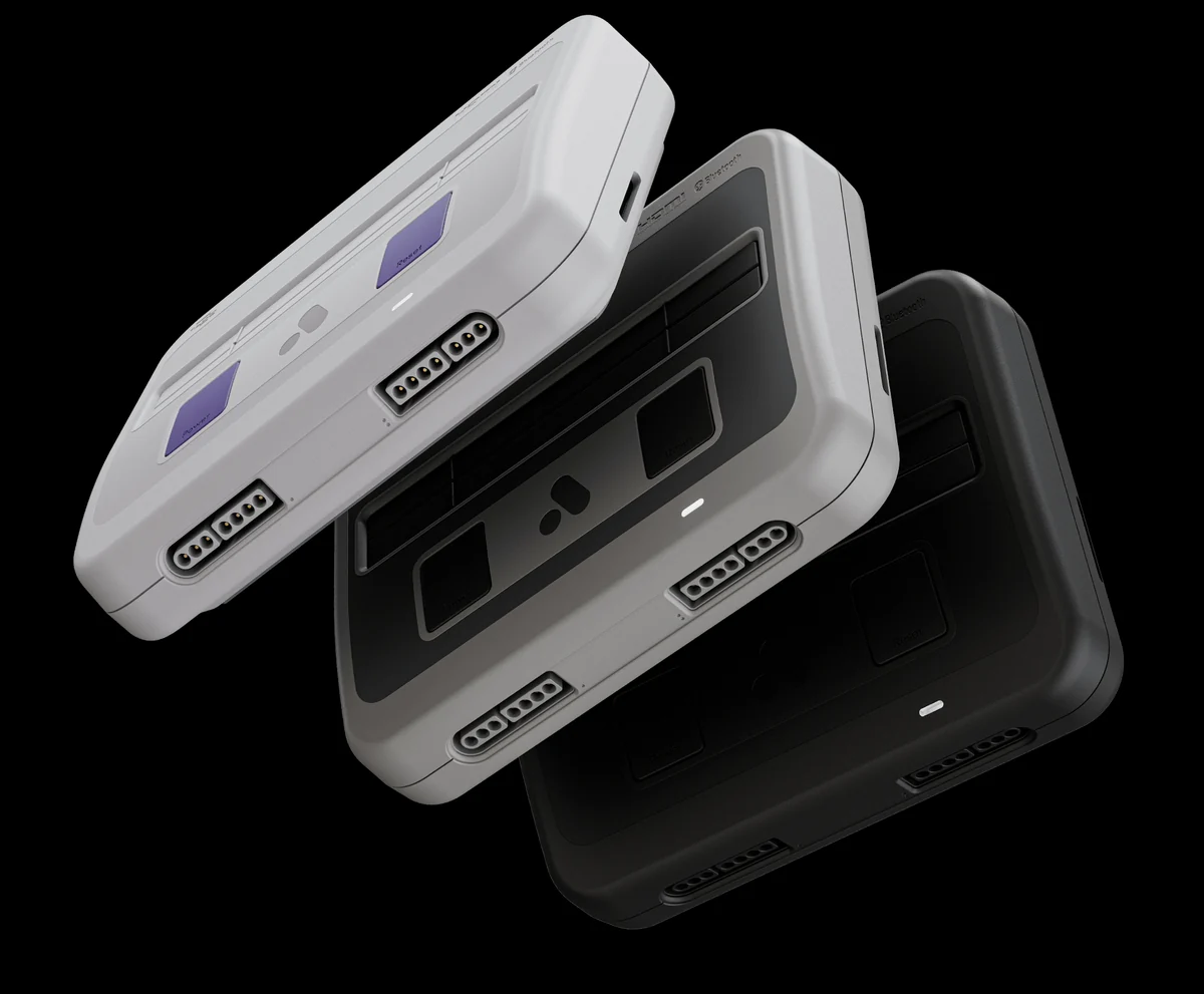 Trois consoles Super Nt+ empilées en biais : une grise claire aux boutons violets, une grise et une noire