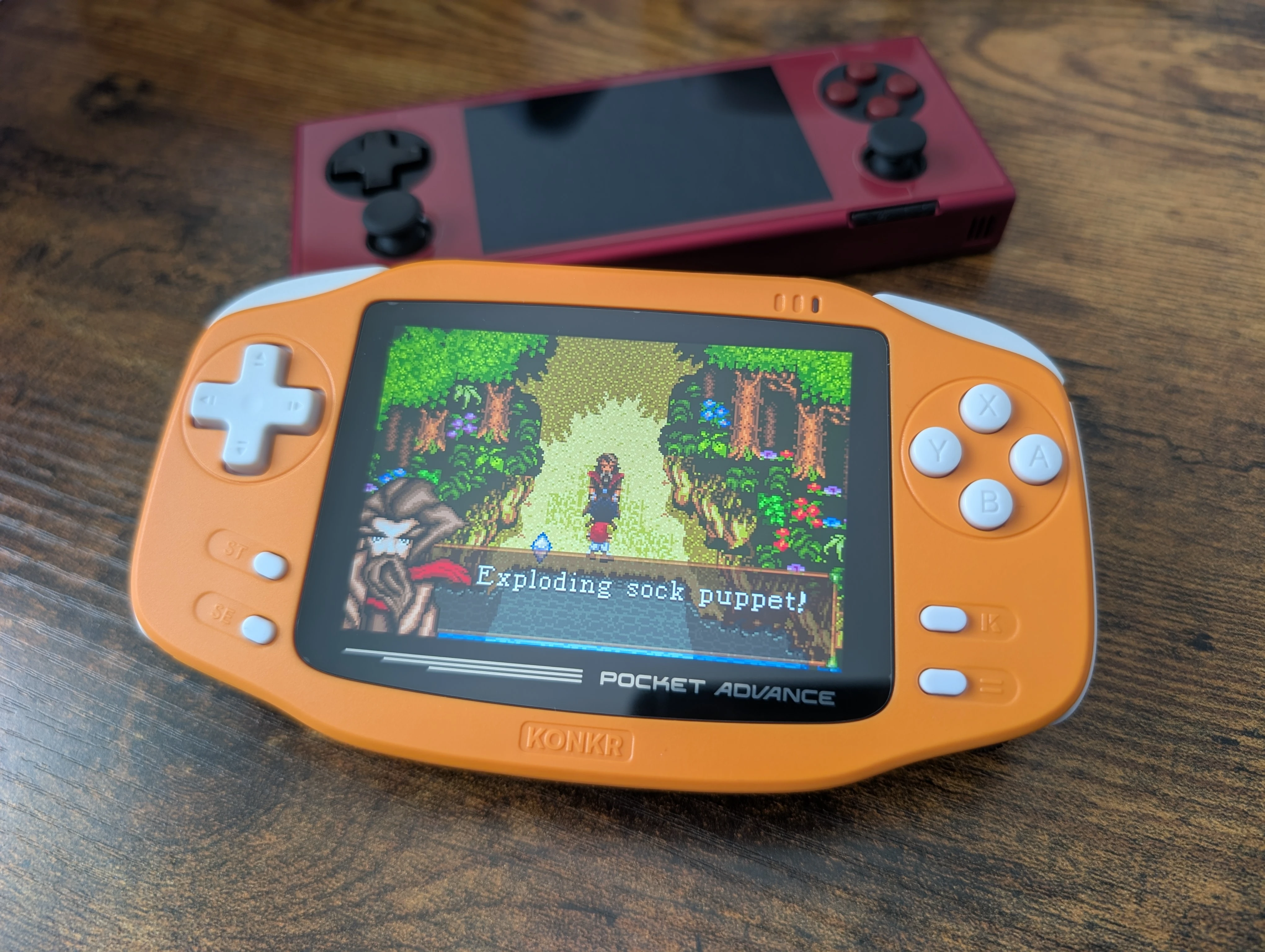 Le KONKR Pocket Advance, console portable orange affichant une scène en pixel art avec le texte « Exploding sock puppet! » à l'écran, posé sur une table en bois devant une autre console portable rouge à l'arrière-plan
