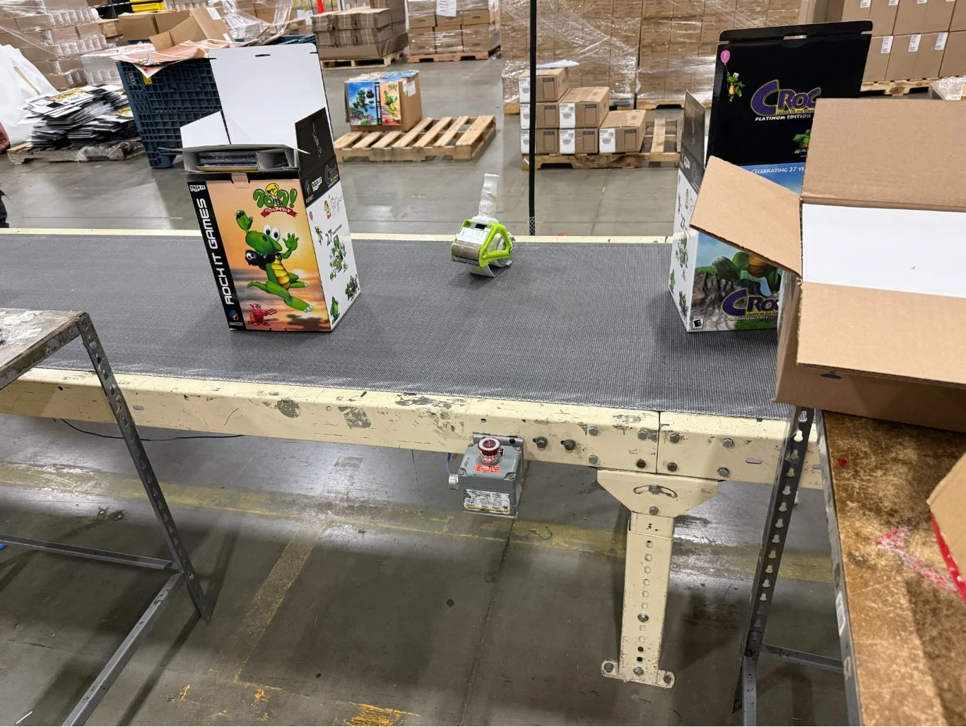 Dans un entrepôt, des boîtes illustrées de Croc posées sur un tapis roulant, à côté de cartons ouverts