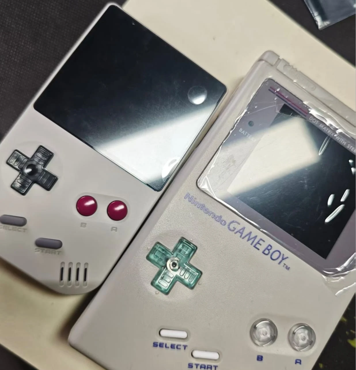 Photo comparant le FPGB Mini, coque blanche avec écran noir, à un Game Boy DMG-01, coque blanc cassé avec écran éteint recouvert de son film plastique et croix directionnelle translucide vert d'eau, posés côte à côte