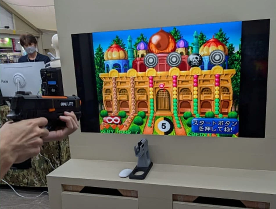 Photo d'une démonstration : un avant-bras à manche camouflage tient un pistolet optique G'AIM'E Lite dirigé vers un écran affichant un jeu de tir de galerie représentant un palais oriental orné de dômes colorés