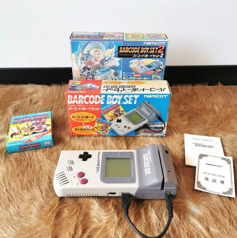Le coffret japonais Namcot Barcode Boy Set : le lecteur de codes-barres relié par câble à une console Game Boy grise posée à côté, devant sa boîte, la boîte du Barcode Boy Set 2 et un jeu Battle Space