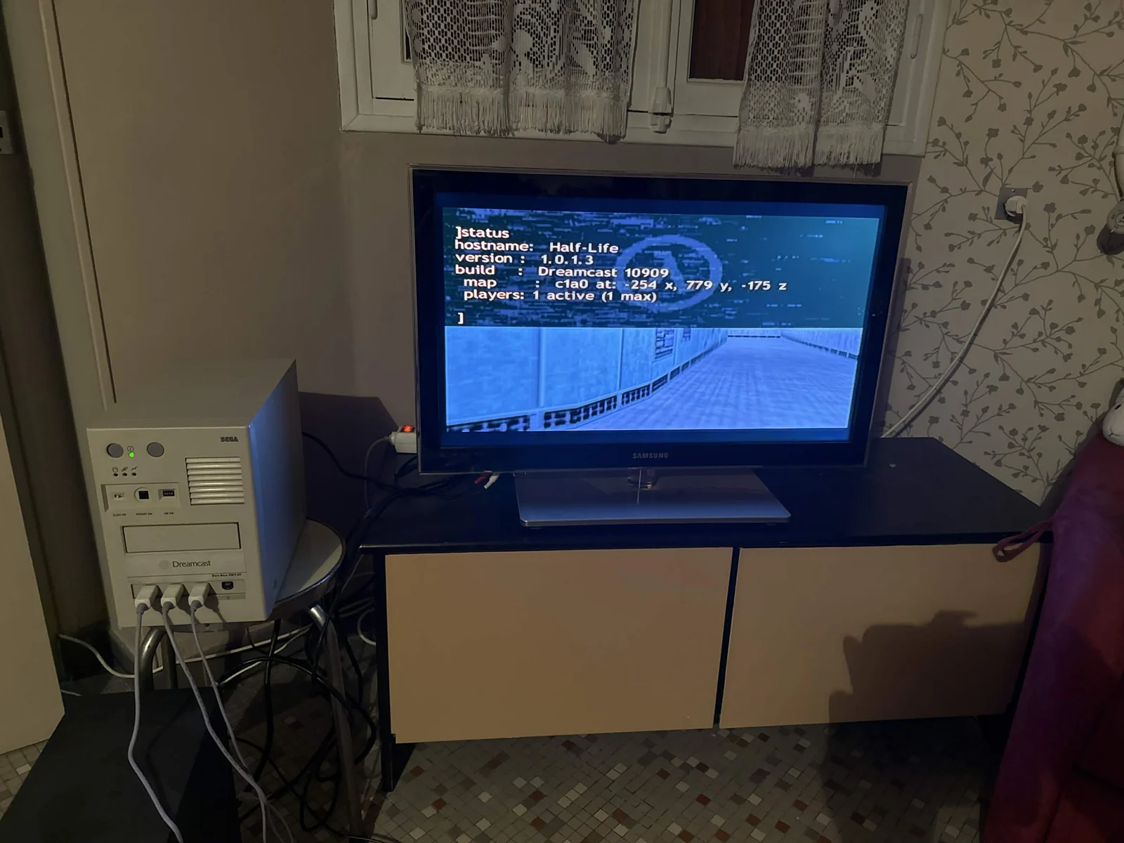 Photo : un kit de développement Dreamcast, boîtier gris marqué Sega et Dreamcast, relié à un téléviseur qui affiche un couloir du jeu et une console de commande indiquant hostname: Half-Life et build: Dreamcast 10909