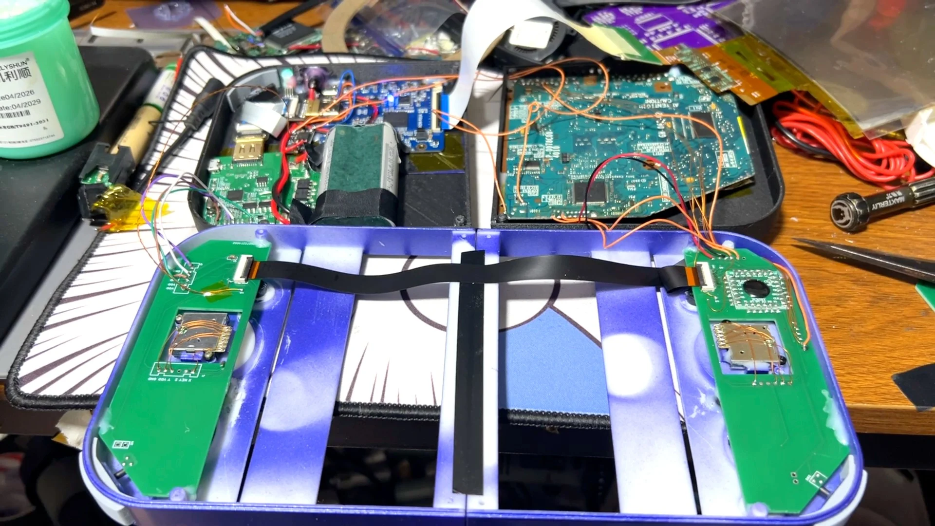 Un boîtier bleu ouvert sur un établi, rempli de circuits et de nappes, entouré de cartes électroniques et d'outils