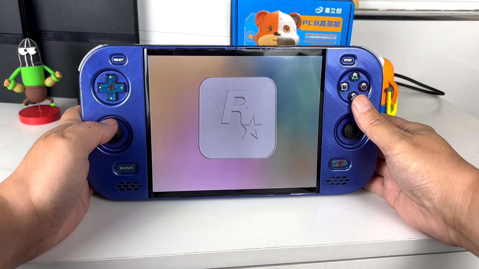 Deux mains tiennent une console portable bleue ouverte, avec croix directionnelle, sticks et boutons PlayStation de chaque côté d'un grand écran qui affiche le logo de Rockstar Games
