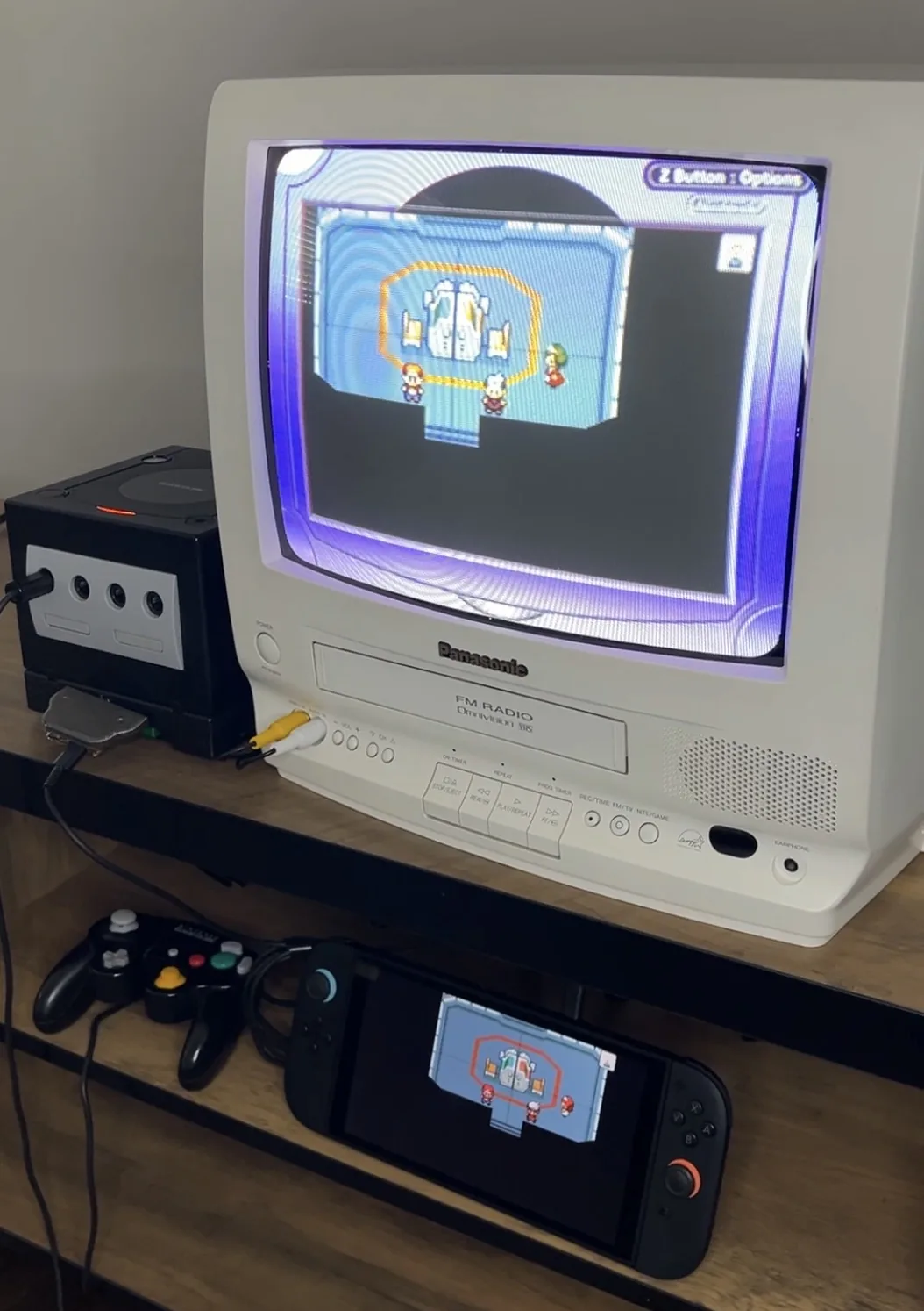 Photo officielle : une GameCube noire posée sur un Game Boy Player, l'adaptateur branché dessous, l'image affichée sur un téléviseur cathodique blanc. Devant, une manette GameCube et une Switch noire qui affiche la même scène