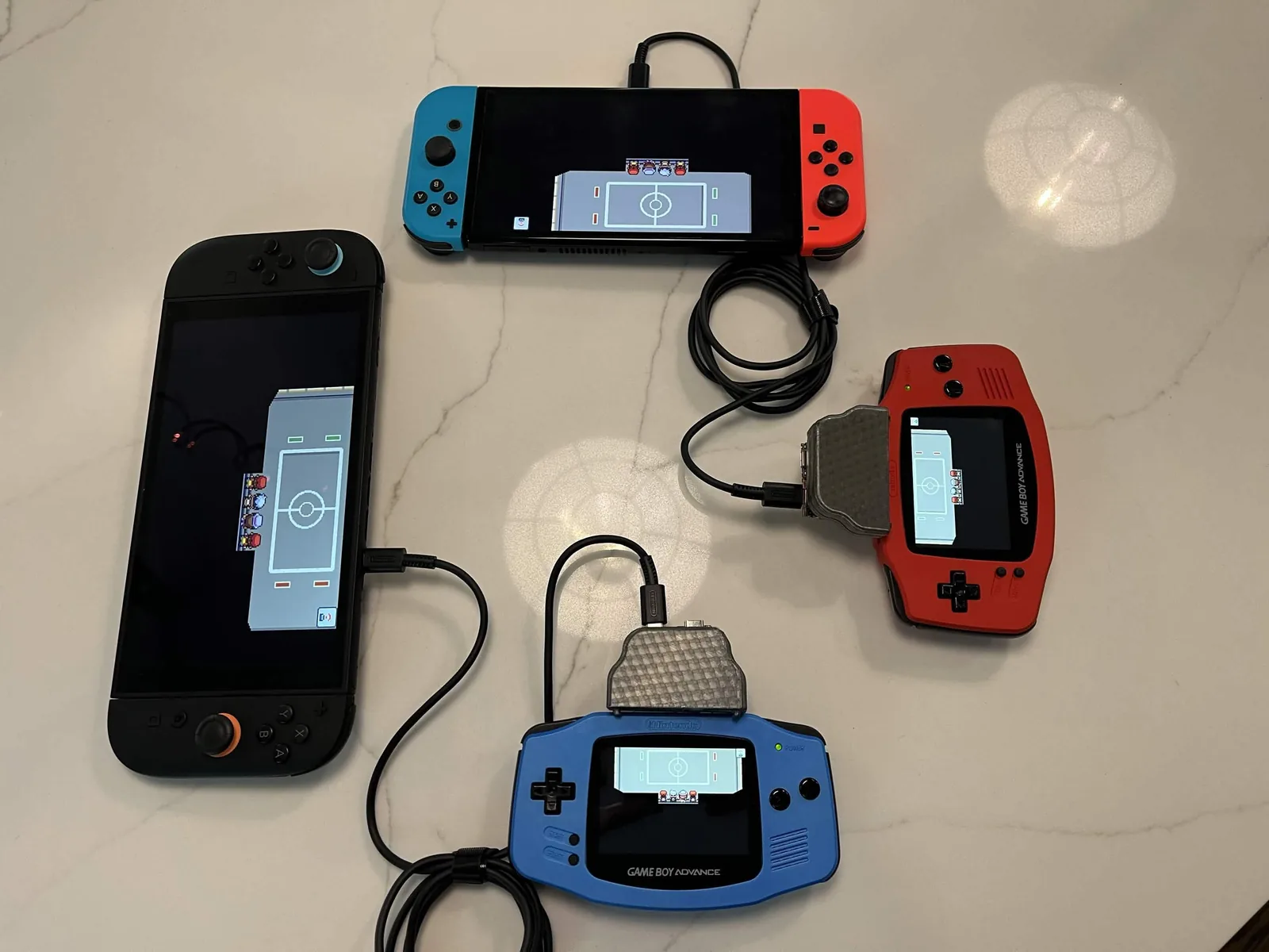 Photo officielle : deux Game Boy Advance, une rouge et une bleue, chacune équipée de l'adaptateur et reliée par un câble à une Switch, l'une aux Joy-Con bleu et rouge, l'autre noire. Les quatre écrans affichent la même salle vue de dessus