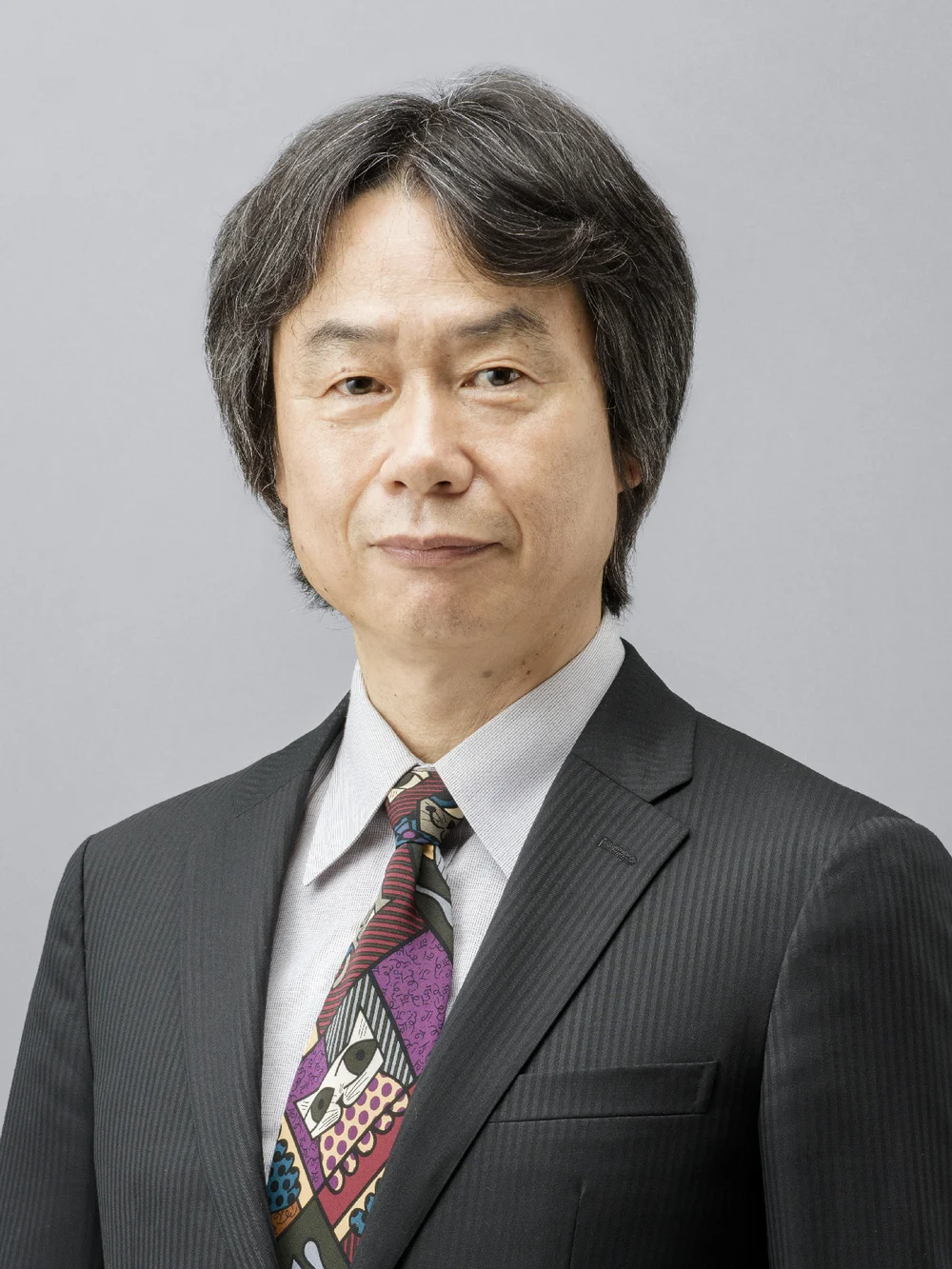 Portrait de Shigeru Miyamoto, cheveux grisonnants, en costume sombre, chemise claire et cravate à motifs, sur fond gris