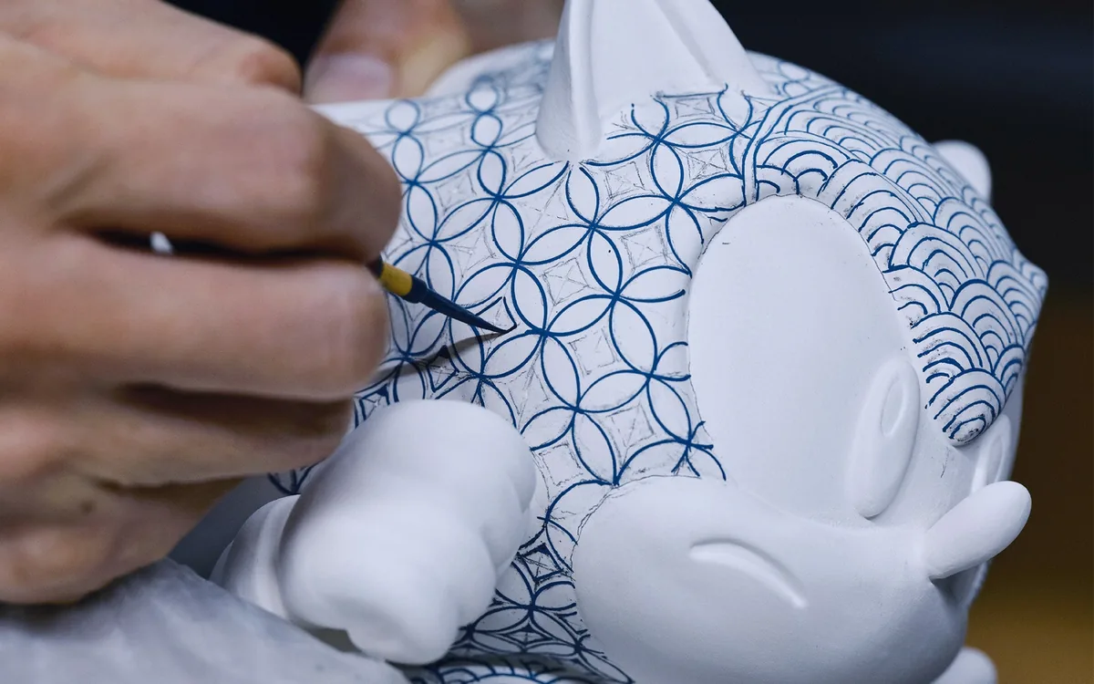 Une main peint au pinceau fin des motifs bleus sur la tête, derrière l'oreille, d'une statue encore en grande partie blanche
