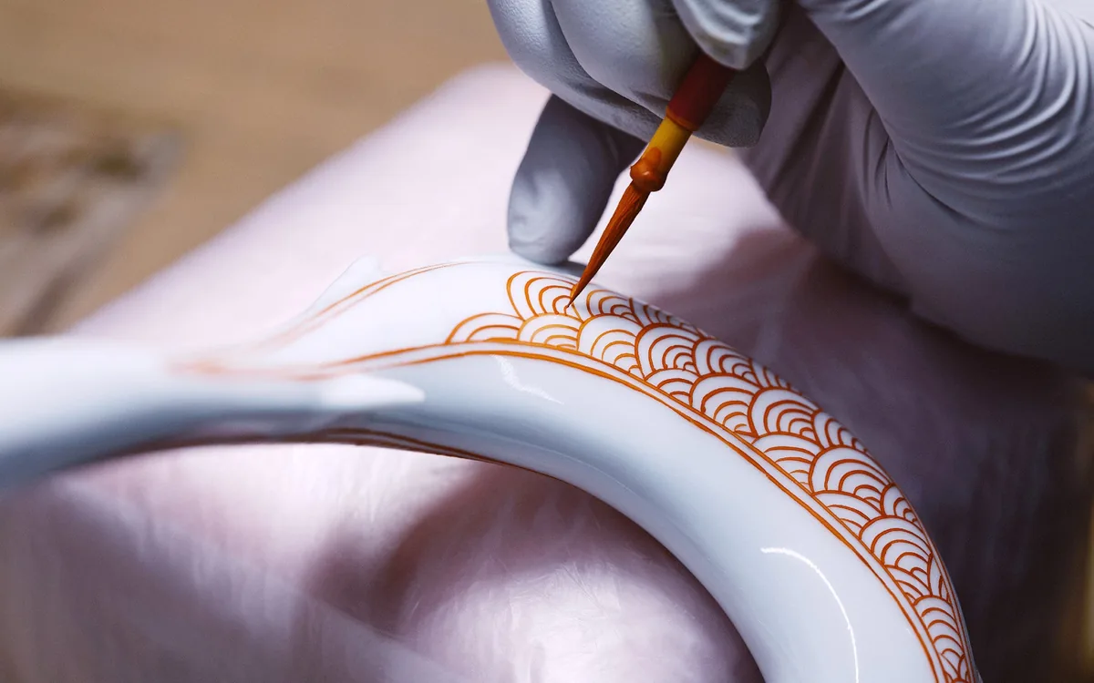 Une main gantée peint au pinceau un motif de vagues orangé sur une boucle de porcelaine blanche