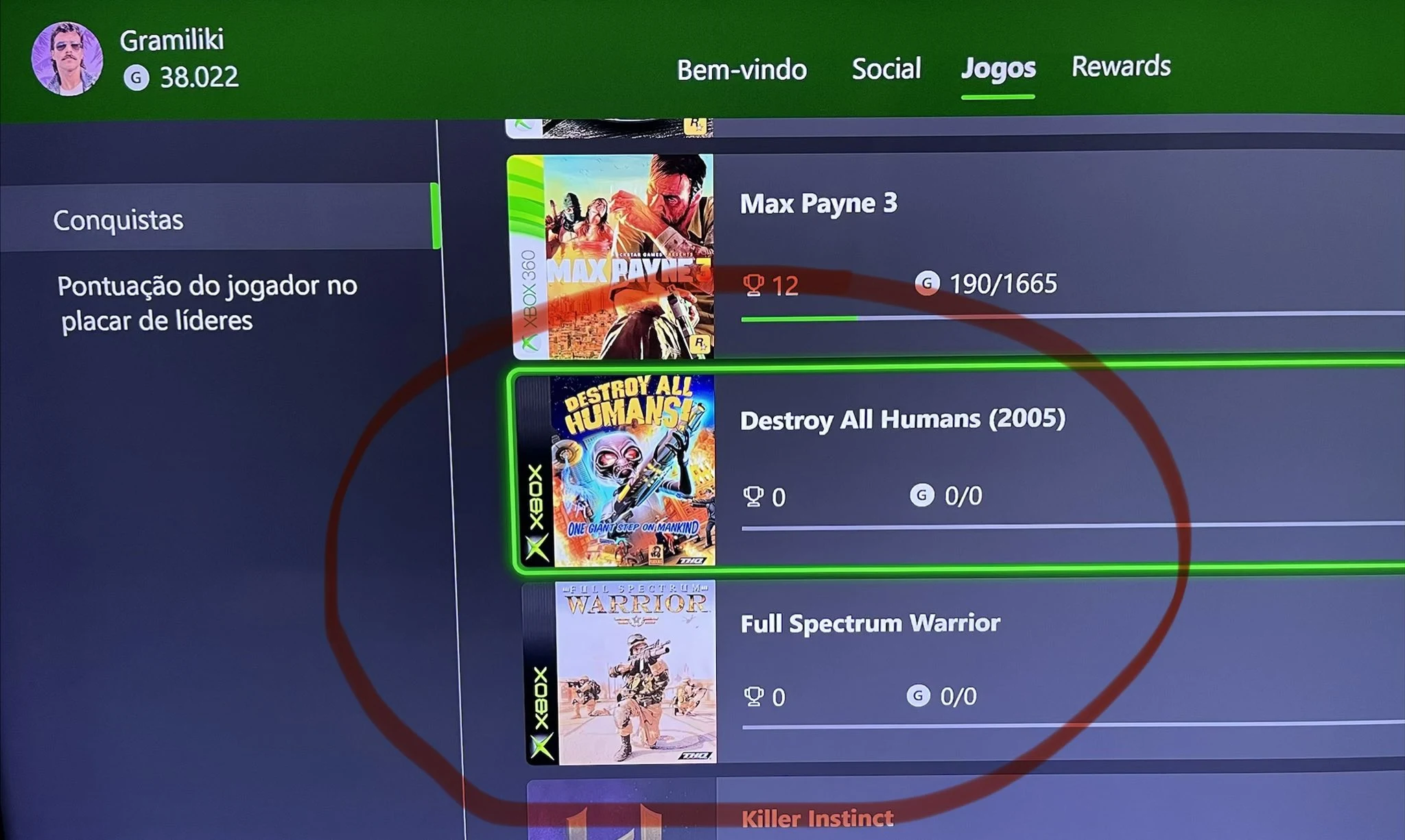 Photo d'un écran affichant la page Conquistas de l'interface Xbox : Max Payne 3 avec 12 succès, puis Destroy All Humans (2005) et Full Spectrum Warrior, marqués du logo Xbox d'origine, à 0 succès et 0 sur 0 points, entourés en rouge
