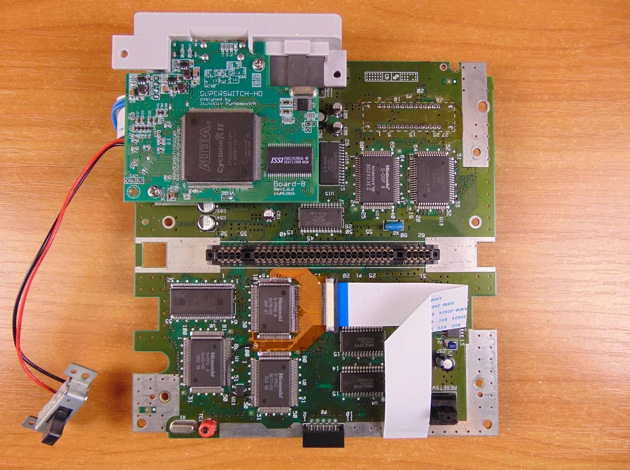 La carte mère d'une Super Nintendo hors de sa coque, avec une carte verte ajoutée près du port de cartouche, une nappe blanche et un adaptateur souple orange