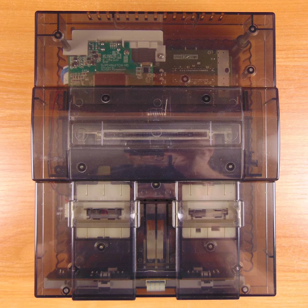 Une Super Nintendo à coque transparente fumée vue de dessus, laissant voir une carte électronique ajoutée à l'arrière, entre la fente à cartouche et les ouïes
