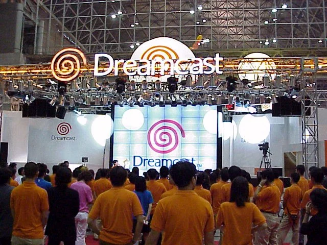 Une grande scène surmontée du logo Dreamcast, devant un public dont la plupart des membres portent des polos orange