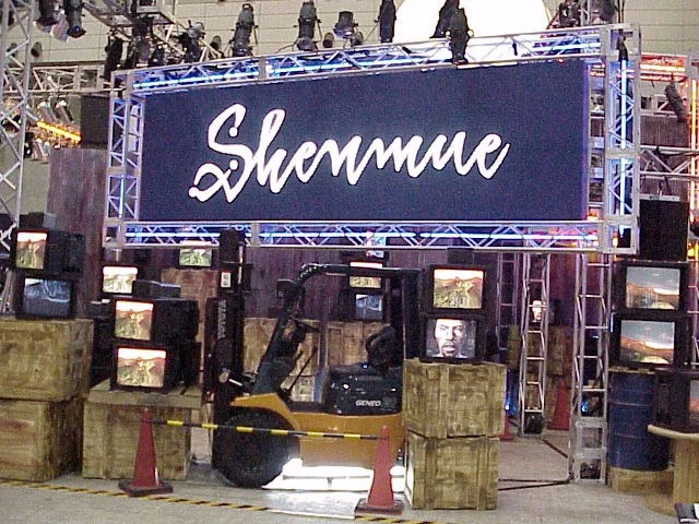 Un stand surmonté d'une enseigne Shenmue, avec un chariot élévateur entouré de caisses et de téléviseurs qui diffusent des images du jeu