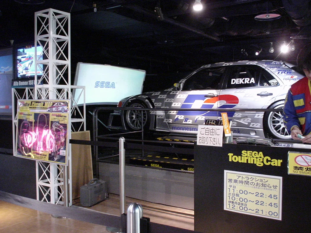 Une voiture de course grandeur nature installée comme une borne d'arcade, devant un écran, avec un panneau Sega Touring Car