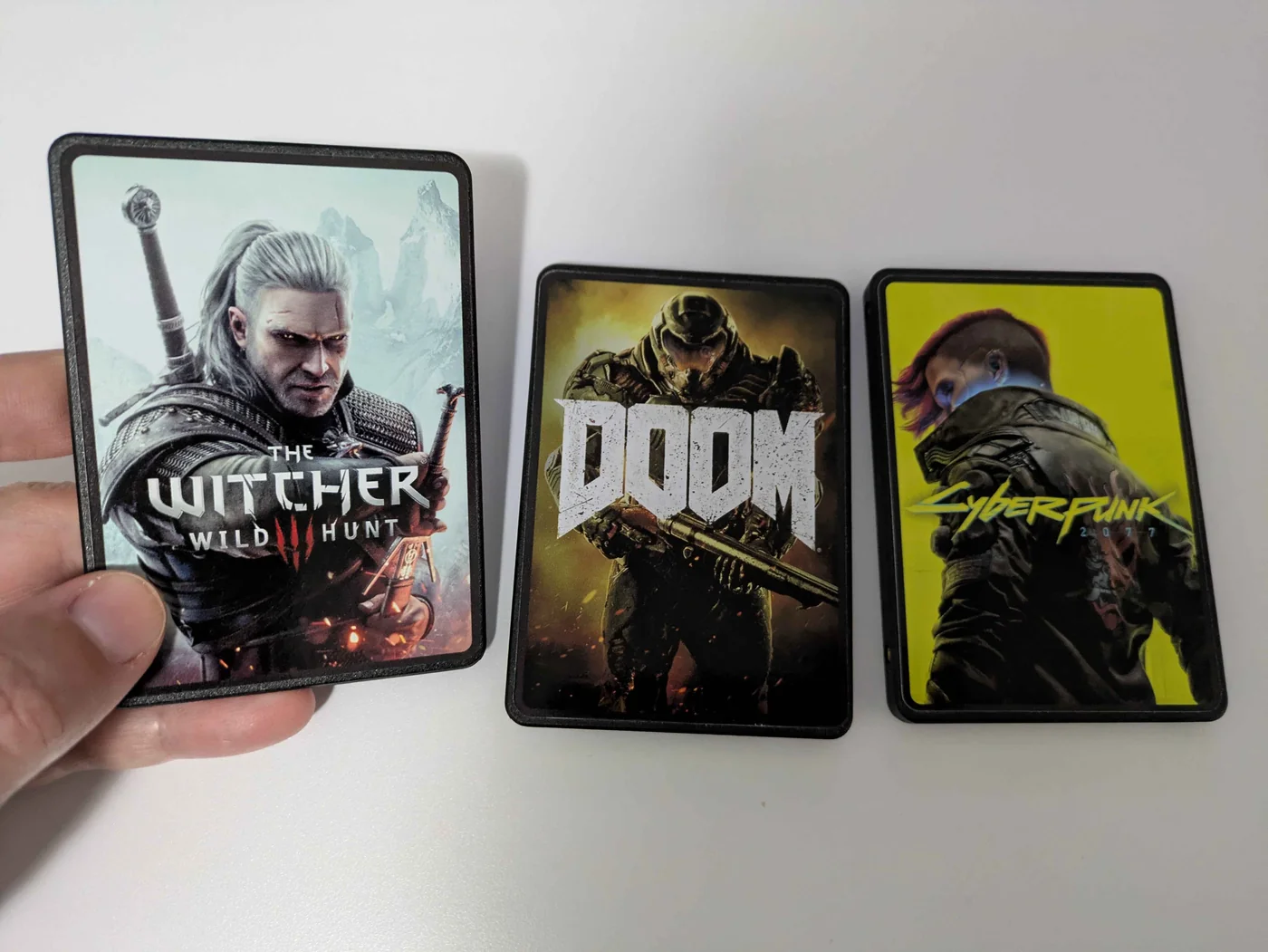 Trois cartouches SSD personnalisées Kazeta tenues à la main, avec les jaquettes de The Witcher 3, Doom et Cyberpunk 2077