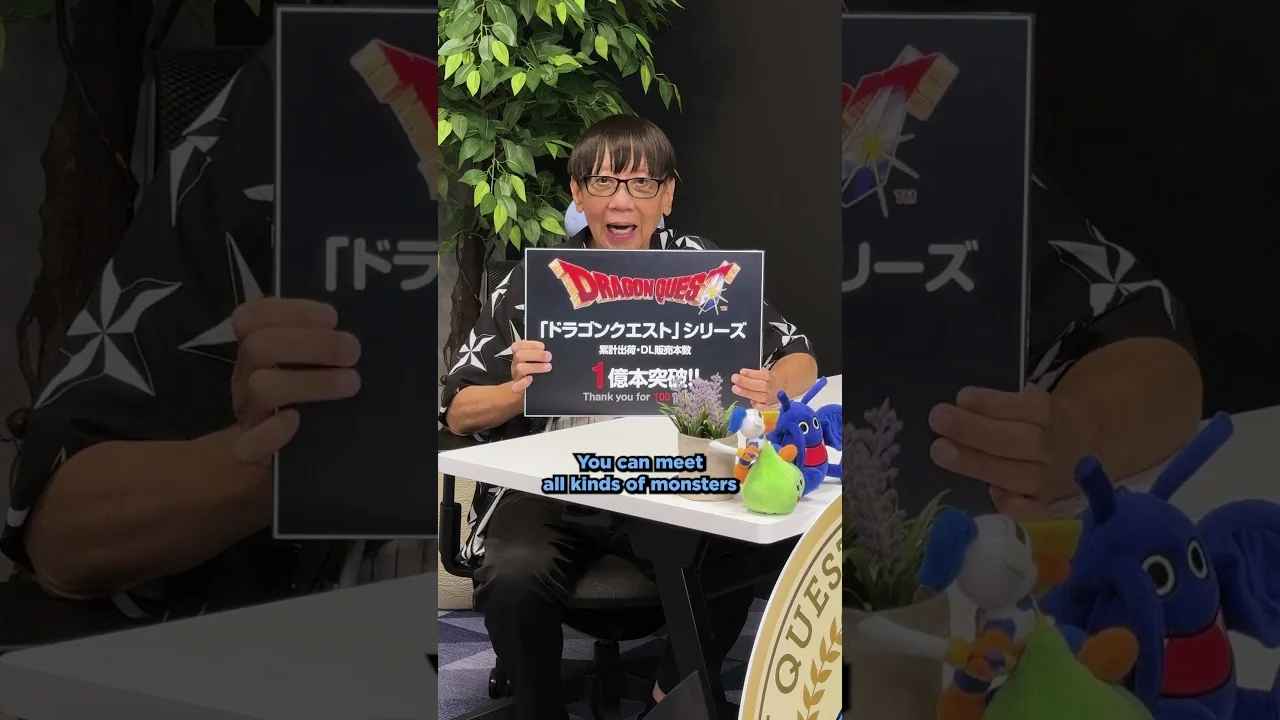 Yuji Horii, assis à un bureau, tient une pancarte en japonais reprenant le logo Dragon Quest et annonçant le cap des 100 millions d'exemplaires, entourée de peluches de monstres de la série dont un Slime bleu
