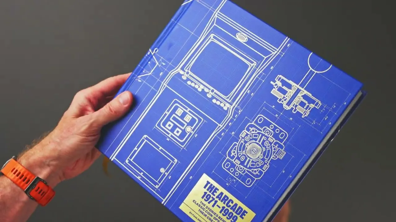 Image de la vidéo officielle : une main tient le livre The Arcade 1971-1999, couverture bleue en plan technique de borne et titre sur cartouche doré